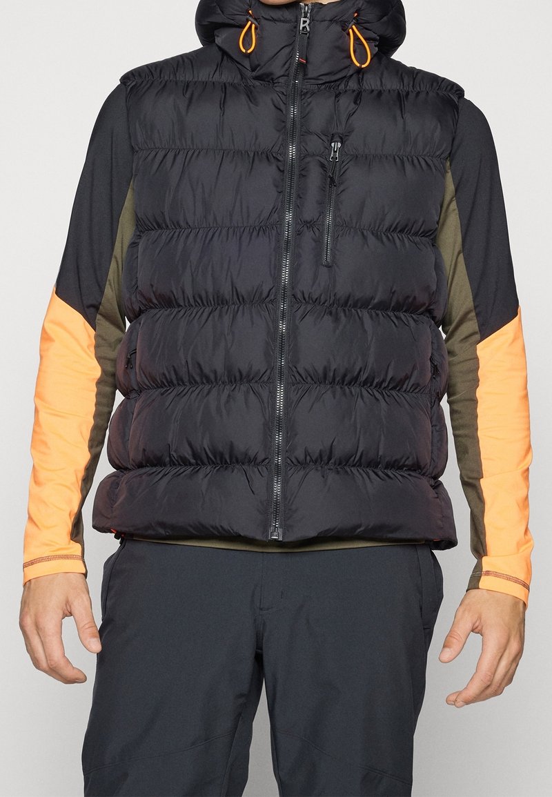 Homme portant une veste matelassée noire avec capuche par-dessus une chemise à manches longues avec des manches orange et vert olive, et un pantalon noir.