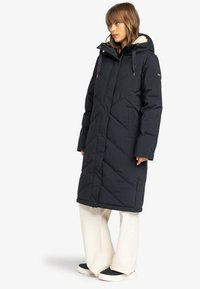 Abrigo largo estilo puffer en azul marino oscuro con diseño acolchado, capucha forrada de pelo y cremalleras verticales. Combinado con pantalones de pierna ancha de color claro.