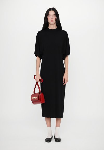 Robe noire avec des manches dolman courtes, associée à un petit sac à main rouge. Le mannequin porte des chaussettes claires et des ballerines noires.