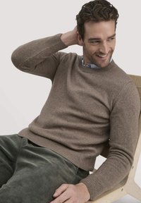 Brauner, gestrickter Pullover mit Rundhalsausschnitt und gerippten Bündchen, kombiniert mit grünen Cordhosen. Model sitzt in einem hellen Holzstuhl.