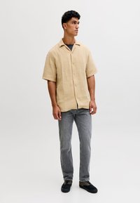 Noor mees seisab, seljas beež lühikeste varrukatega tekstuuriga särk, hallid teksad ja mustad tossud, taust on ühtlane.