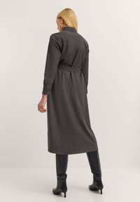 Langes, dunkelgraues Kleid mit einem Kragen, langen Ärmeln und einem taillierten Bund. Es hat Knopfmanschetten und eine gerade Silhouette, kombiniert mit hohen schwarzen Stiefeln.