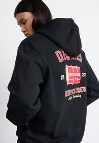 Felpa nera con cappuccio che presenta una grafica sulla schiena con testo rosso e beige. Include un design a bott barrel e la scritta "SERVICE CREW". Tessuto morbido, vestibilità rilassata.