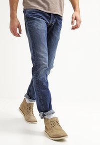 Hombre caminando, vistiendo jeans azules arremangados, calcetines grises y botas de ante marrón claro con cordones, sobre fondo blanco.