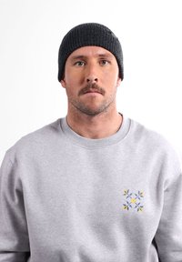 Sweatshirt gris avec un col rond et un petit motif floral en bleu et jaune sur la poitrine gauche. Bonnet en tricot noir sur la tête.