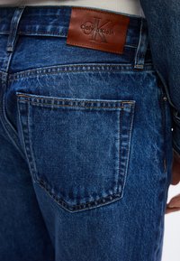 Κοντινό πλάνο πίσω τσέπης μπλε τζιν με καφέ δερμάτινη ετικέτα μάρκας Calvin Klein πάνω από την τσέπη.