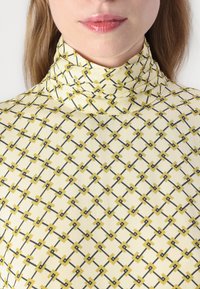 Tricou cu guler înalt și mâneci lungi din țesătură galben deschis, cu un model geometric în nuanțe de negru și galben, având o textură netedă.