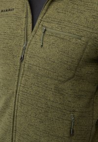 Giacca in pile verde con tessuto testurizzato, chiusura con zip sul davanti e tasca sul petto. Presenta un collo alto e un sottile motivo a macchie scure.