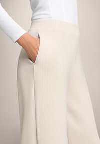 Bež hlače širokih nogavica s vertikalnim trakama. Imaju bočne džepove i glatki pojas. Tkanina izgleda teksturirano i krojeno.