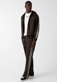 Brązowy dres w wzory z rozpinaną bluzą z kapturem, białe akcenty i paski po bokach. Połączony z białym t-shirtem z logo i czarnymi trampkami.