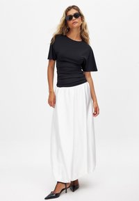 Juoda trumpa rankovėmis palaidinė su susiraukšlėta tekstūra, derinama su plaukuojančia balta maksi sijonu. Modelis dėvi juodus aštriakampius batelius ir saulės akinius.