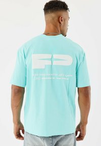 Camiseta de manga corta color turquesa, con un gran texto gráfico blanco en la parte posterior. Corte casual con una textura suave y diseño de cuello redondo.