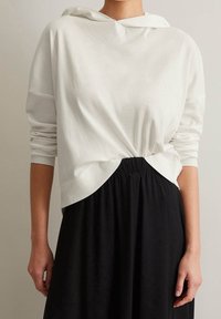 Maglione bianco con cappuccio e maniche lunghe, caratterizzato da una finitura testurizzata e un dettaglio annodato sul davanti, abbinato a una gonna nera.