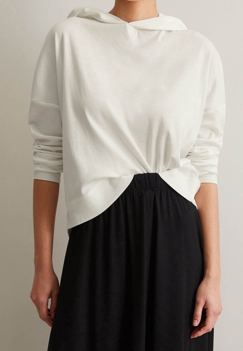 Maglione bianco con cappuccio e maniche lunghe, caratterizzato da una finitura testurizzata e un dettaglio annodato sul davanti, abbinato a una gonna nera.