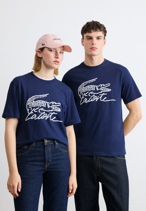 Junger Mann und Frau tragen marineblaue Lacoste-T-Shirts mit weißem Krokodil-Logo; die Frau trägt außerdem eine pinke Lacoste-Kappe und blaue Jeans.