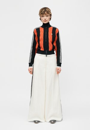 O persoană stă cu fața spre înainte, purtând o jachetă cu fermoar în dungi negre și portocalii, pantaloni albi largi și pantofi argintii.