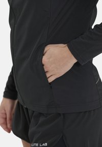 Schwarze Sportjacke mit langen Ärmeln und einer Reißverschlusstasche, kombiniert mit schwarzen Shorts, die ein dezentes Logodesign aufweisen. Glattes, leichtes Material.