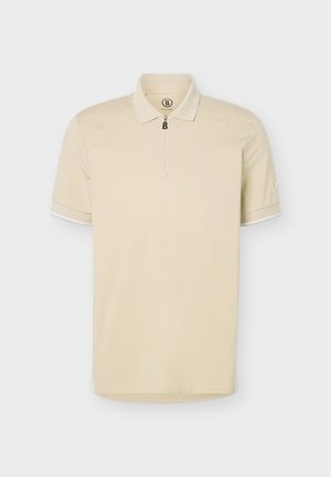 Hombre de pie mirando hacia adelante con polo beige de manga corta, pantalones negros con cinturón y zapatillas blancas sobre fondo claro.