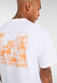 Bílá bavlněná tričko s oranžovým grafickým designem dvou kovbojů, koní a textem "VĚČNÝ SPOLUBYDLÍCÍ" na zádech.