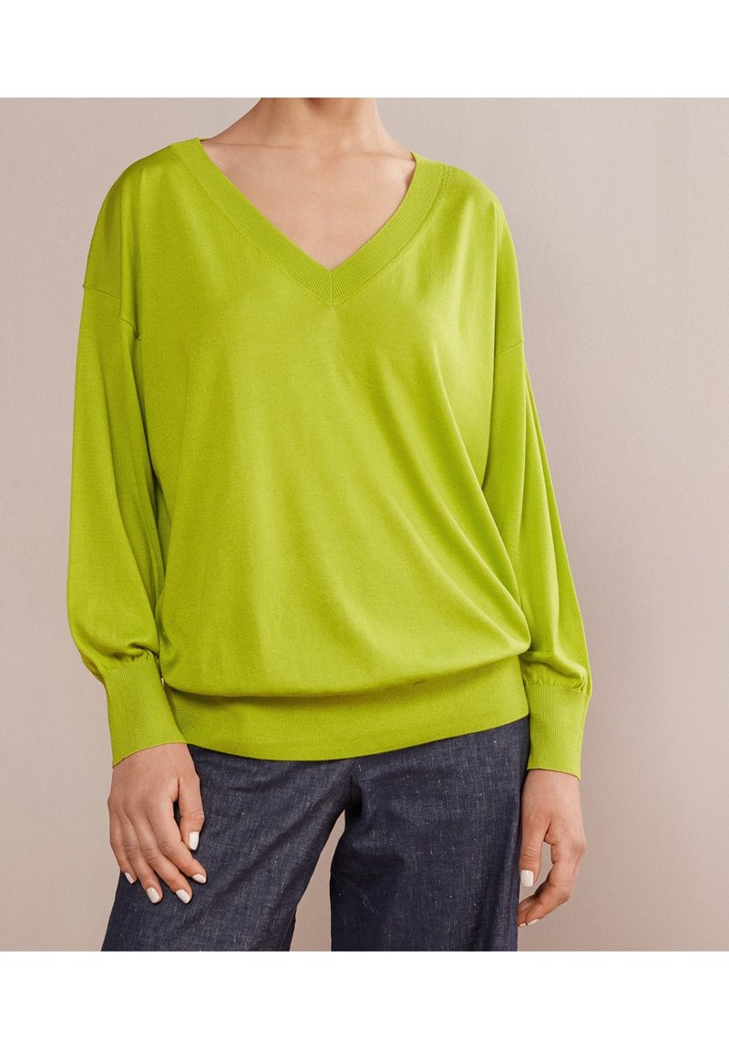 Personne portant un pull vert citron à col en V à manches longues et un pantalon bleu foncé, une main reposant sur la cuisse sur un fond uni.