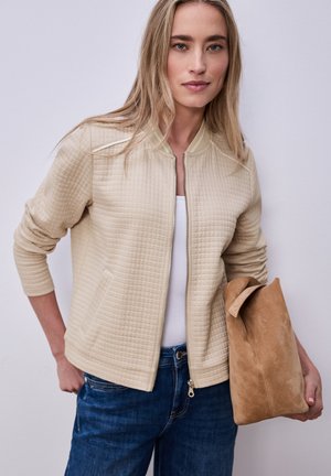 Femme blonde portant une veste matelassée beige sur un haut blanc et un jean bleu, tenant un sac à main en daim beige sur un fond uni.