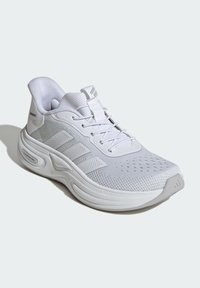 Bijela Adidas tenisica za trčanje s mrežastim gornjim dijelom, podstavljenim potplatima, vezanjem na vezice i tri pruge sa strane, postavljena pod kutom lijevo na svijetloj pozadini.