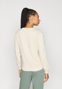 Kremfarget genser med lange ermer og ribbet mansjetter, med en vid hals og myk tekstur. Modellen har på seg lys grønne bukser.