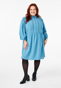 Šviesiai mėlyna, kelių ilgio suknelė su pūstomis rankovėmis ir aukštu apykakle. Yra priekiniai mygtukai ir nėrinių detalės aplink kūną.