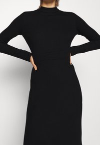 Robe noire à manches longues avec un col montant, une coupe ajustée, une texture lisse et une couture de taille définie.