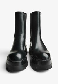 Ghete negre de gleznă din piele cu finisaj lucios, panouri elastice laterale și tălpi groase din cauciuc negru cu crampoane zimțate pentru aderență.