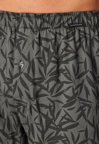 Boxers en tissu vert foncé avec un motif de feuilles noires, taille élastique, bouton de fermeture et une petite étiquette de marque.