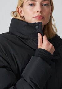 Giacca a vento nera con colletto alto, polsini a costine e dettagli con chiusura a scatto. Texture liscia e silhouette strutturata.