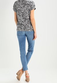 Šedý top s květinovým vzorem a krátkým rukávem, kombinovaný se světle modrými skinny džíny. Béžové sandály na vysokém podpatku s páskami kolem kotníků. Jednoduché pozadí.