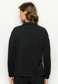 Donna con capelli castani mossi, indossa una maglietta nera a maniche lunghe e pantaloni neri, in piedi di fronte a un muro bianco.