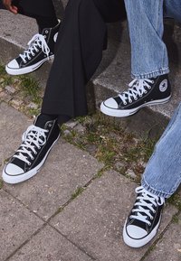 Två personer sitter på betongtrappor och har på sig svarta och vita högskurna Converse-sneakers, en med svarta byxor och den andra med blå jeans.