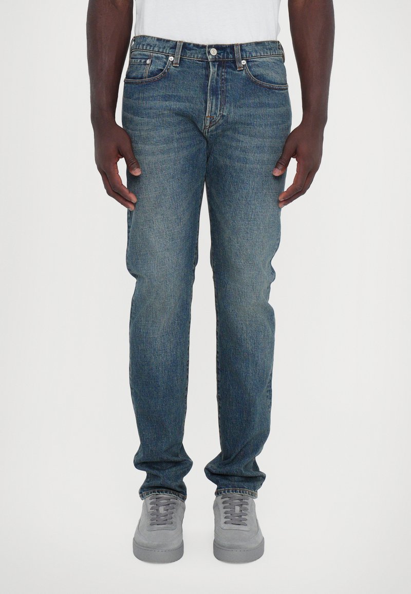 Jean en denim bleu foncé avec une coupe droite, design à cinq poches et un système de fermeture à bouton à la taille. Porté avec des baskets grises.