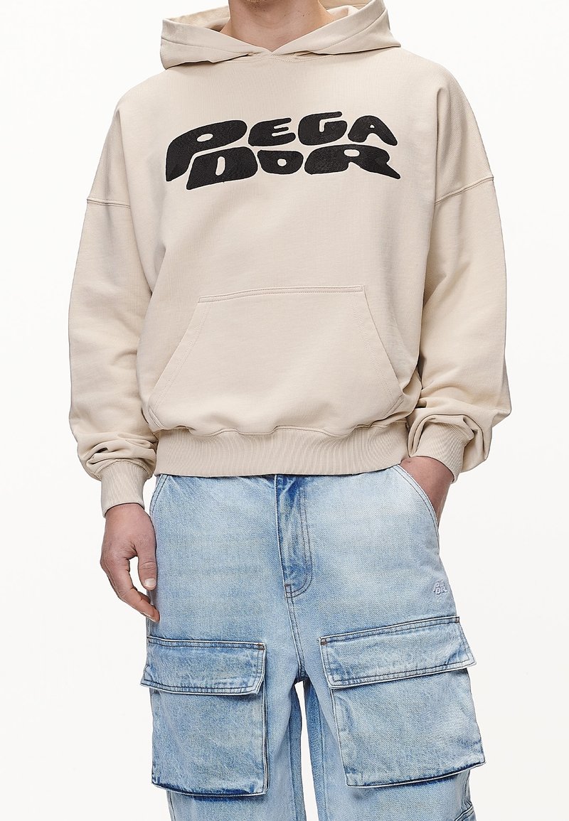 Sudadera con capucha beige con un estampado gráfico negro, con un bolsillo frontal y puños de canalé, combinada con pantalones cargo azul claro.
