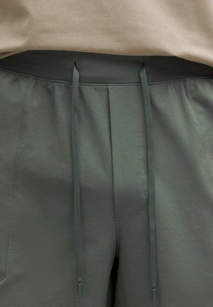 Nahaufnahme von grauen Jogginghosen mit strukturiertem Stoff und einem beigen Hemd, das an der Taille gesteckt ist.