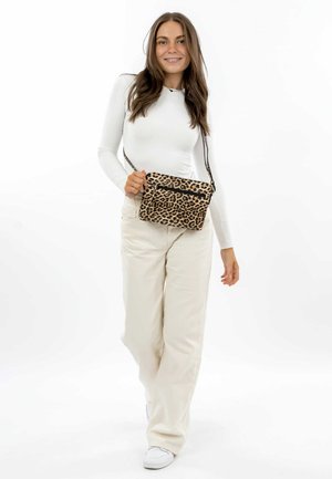 Leopard print crossbody bag with two zippered pockets, soft material, and adjustable strap, paired with a white long-sleeve top and beige pants.