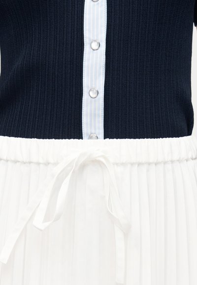 Μπλε ναυτικό ριμπ τοπ με κάθετη ρίγα κουμπιών και ασημένια κουμπιά, σετ με λευκή πλισέ φούστα που διαθέτει κορδέλα μέσης δεμένη.
