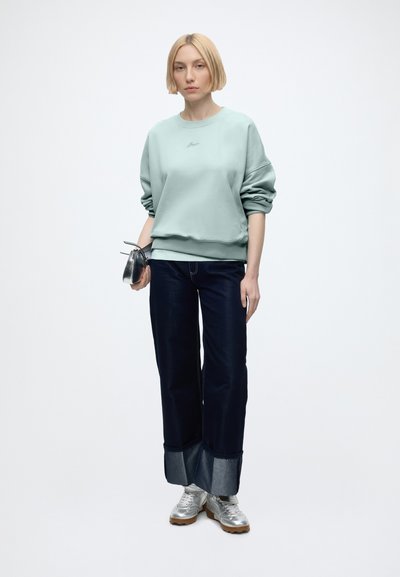 Femme aux cheveux blonds courts portant un sweat-shirt bleu clair, un jean large sombre avec des revers, des baskets argentées et tenant un petit sac argenté.