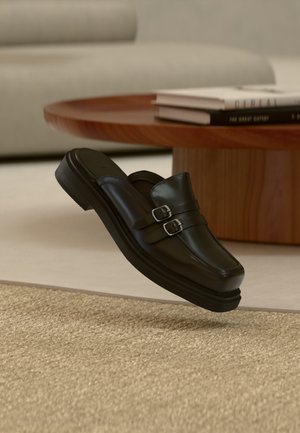 Mustast nahast slip-on jalanõu kahekordsete hõbedaste pandlatega, kujutatud õhus tekstuurse beežika vaiba kohal, puidust kohvilauda ja raamatute lähedal.