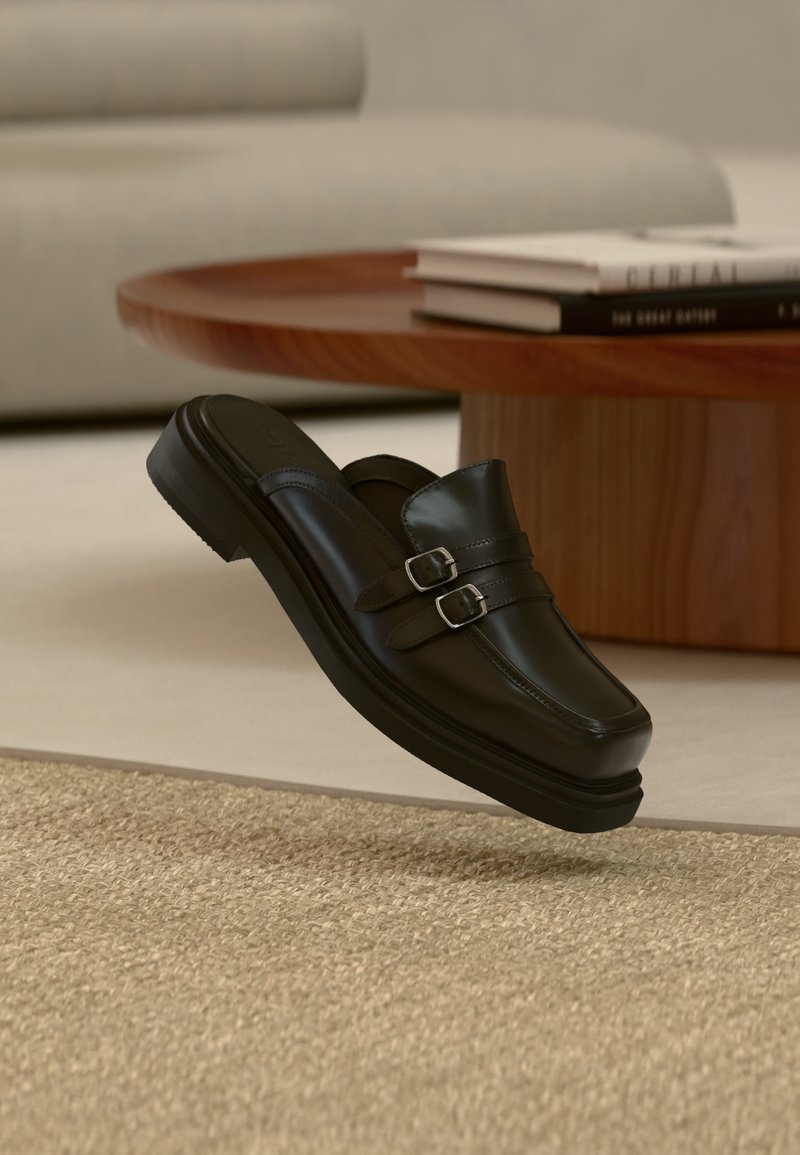 Μαύρο δερμάτινο παπούτσι χωρίς κορδόνια με διπλές ασημένιες πόρπες, αιωρούμενο πάνω από μία υφασμάτινη μπεζ μοκέτα, κοντά σε ξύλινο τραπεζάκι σαλονιού και βιβλία.