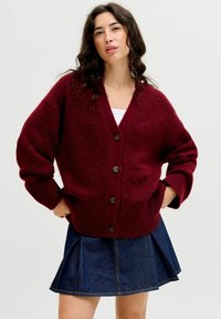 Burgunderfarbener Strickcardigan mit V-Ausschnitt, übergroßen Ärmeln, braunen Knöpfen und strukturierter Oberfläche. Kombiniert mit einem Jeansrock.