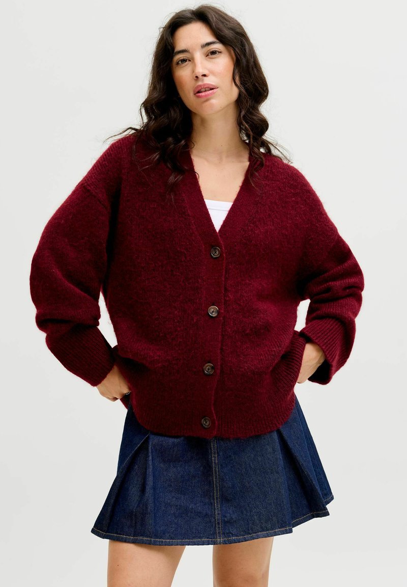 Burgunderfarbener Strickcardigan mit V-Ausschnitt, übergroßen Ärmeln, braunen Knöpfen und strukturierter Oberfläche. Kombiniert mit einem Jeansrock.