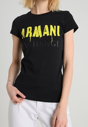 Woman with short blonde hair wearing a black Armani Exchange T-shirt with yellow and gray text, paired with white pants against a plain background.