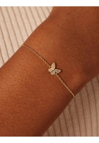 Gouden kettingarmband met een vlinderbedel versierd met heldere stenen, geplaatst op een bruine huidskleur. Eenvoudig, delicaat ontwerp.