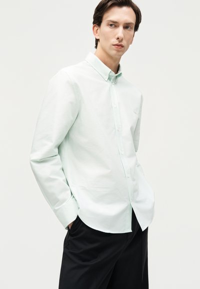 Νέος άνδρας που φοράει ανοιχτοπράσινο πουκάμισο με κουμπιά και μαύρο παντελόνι, στέκεται με τα χέρια στις τσέπες, κοιτάζοντας στο πλάι.