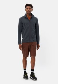 Giacca in pile zipata di un grigio scuro con colletto stand-up, abbinata a pantaloni cargo marroni. Sneakers nere con dettagli gialli. Sfondo bianco.