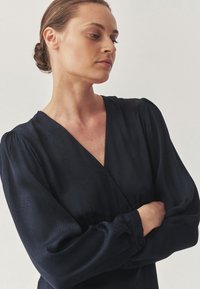 Blusa azul marino con escote en V y mangas largas abullonadas, que presenta un sutil patrón texturizado y detalle de botones en los puños.